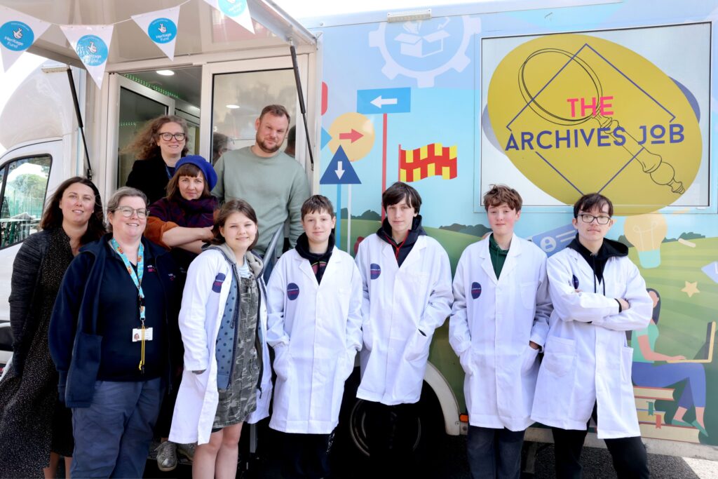 Several adults and several children wearing white lab coats pose in front of a van marked with a gold logo that reads: 'The Archives Job'.