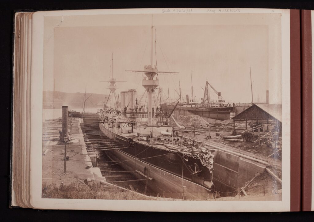 A sepia photograph of a docked ship with two masts. In the background, there are other ships and some wooden structures.
