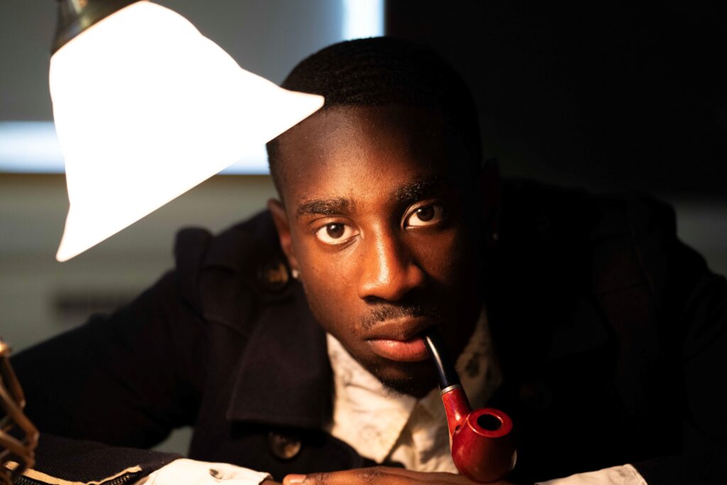 A man smoking a pipe stares into the camera, his face half illuminated by a lamp next to his head.