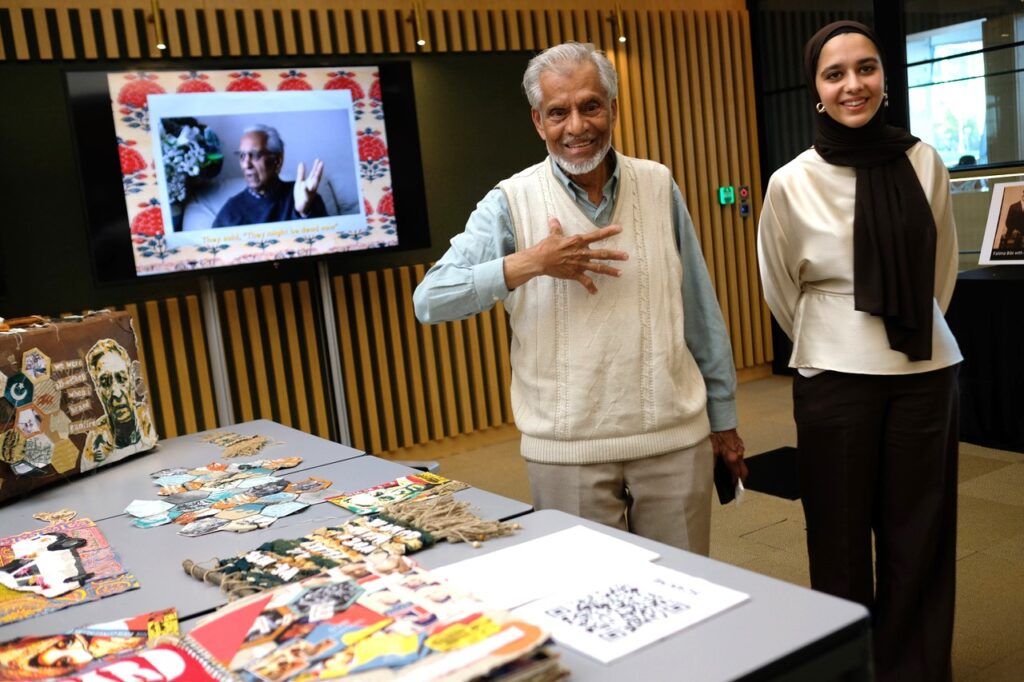 Two people stand by a table of brightly-coloured materials from the project, with a digital screen showing a photo behind them