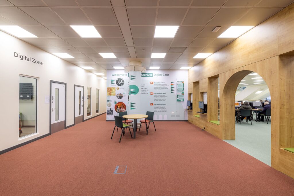 A large room with a red carpet and white walls. 'Digital Zone' is printed on the wall, a table and chairs sit in the centre of the room, in front of an exhibition board. Next door, through an archway, several people can be seen working on computers.