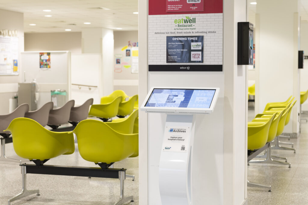 A touch-screen display right next to a waiting area in a hospital filled with yellow seats