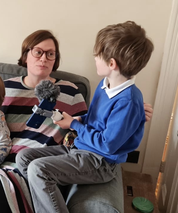 A boy in blue school uniform sitting on the edge of a sofa holding out a microphone to interview a woman