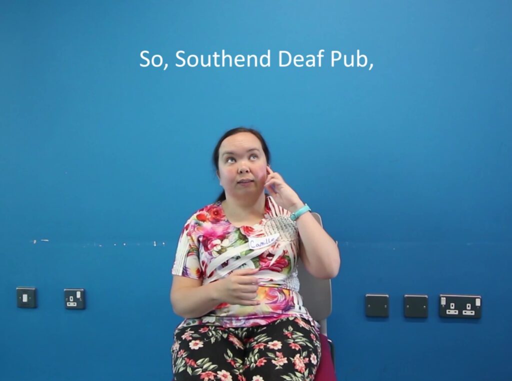A screenshot of a video where a woman is sitting on a chair in front of a blue background and using sign language, with captions written above her