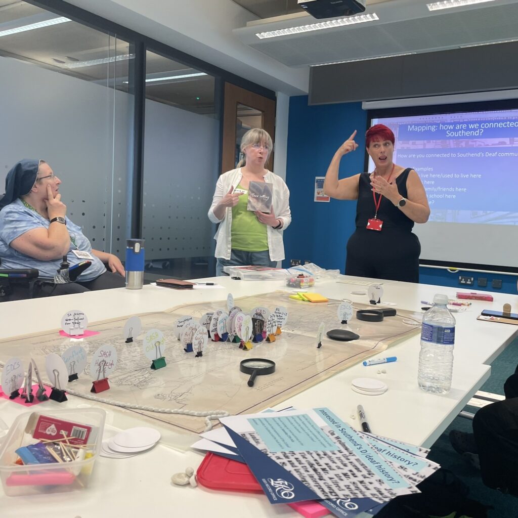 A workshop being led in sign language, with participants sitting round a table that has a map with tokens on it