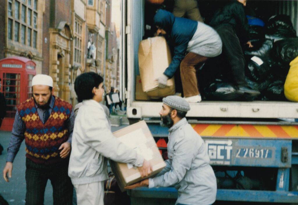 A group of six men, of varying ages, unloading a lorry filled with cardboard boxes and black plastic bags onto a wide pavement.