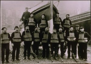 15 men pose for a photo in front of an RNLI boat. All are wearing dark dress, and thirteen of them are wearing contemporary life jackets.