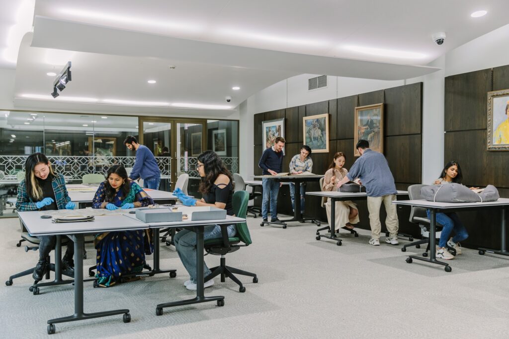 Tables of various sizes in a well-lit room, with a few hanging artworks along one wall. Nine people are in the room, standing or sitting at tables, studying original documents.