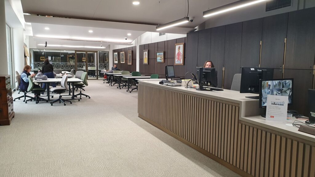 A large open-plan room, with wooden panelling along one wall, from which are hung six artworks. There is a long reception desk in the foreground, and a series of tables in the background.