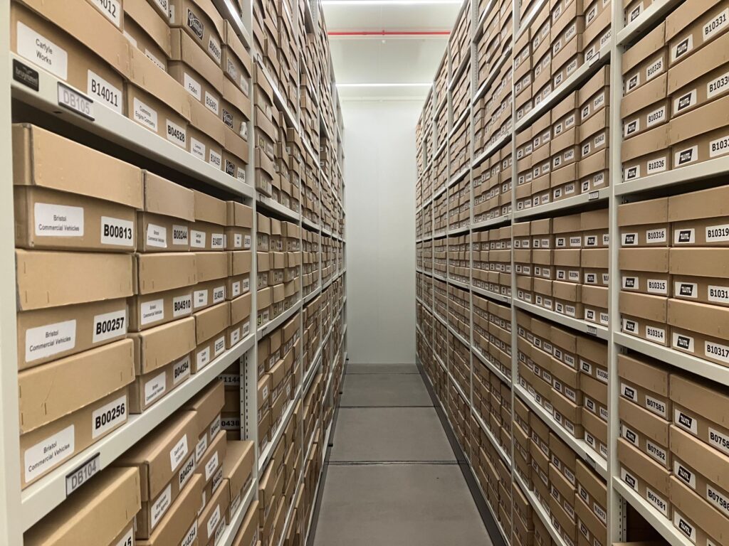 View down an archive storage corridor - floor-to-ceiling racking is full of numbered cardboard boxes, all uniform in size.