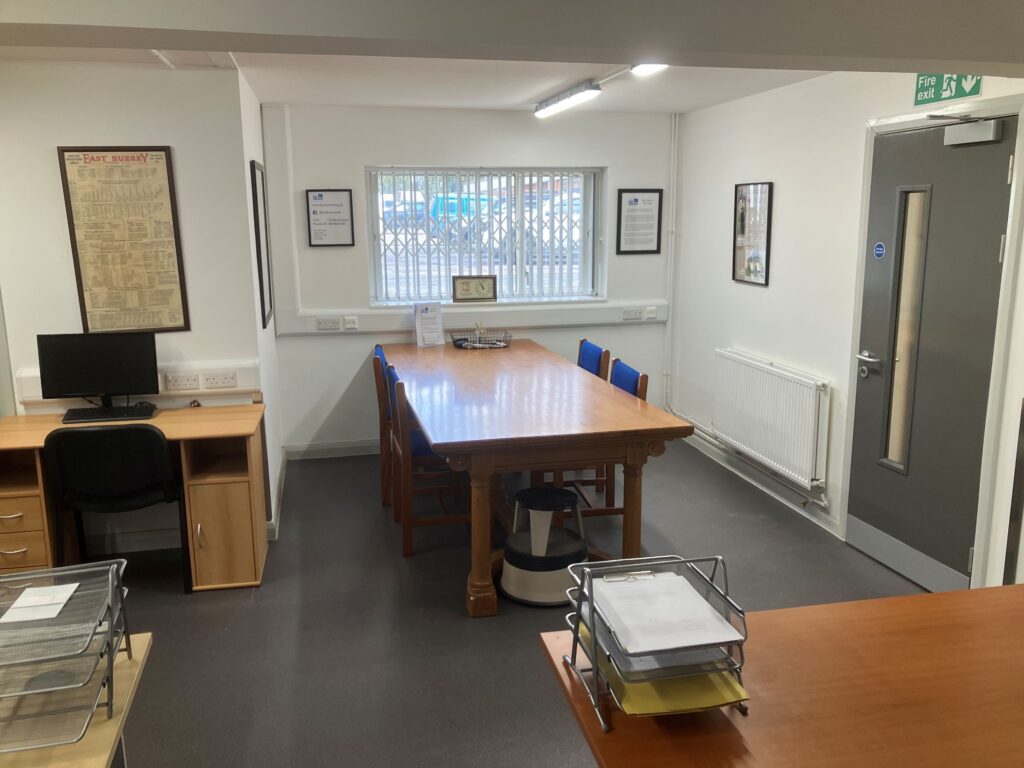 A white-walled room with a computer desk placed against one wall, and a large wooden table jutting out into the room. The wooden table has two chairs on either side.