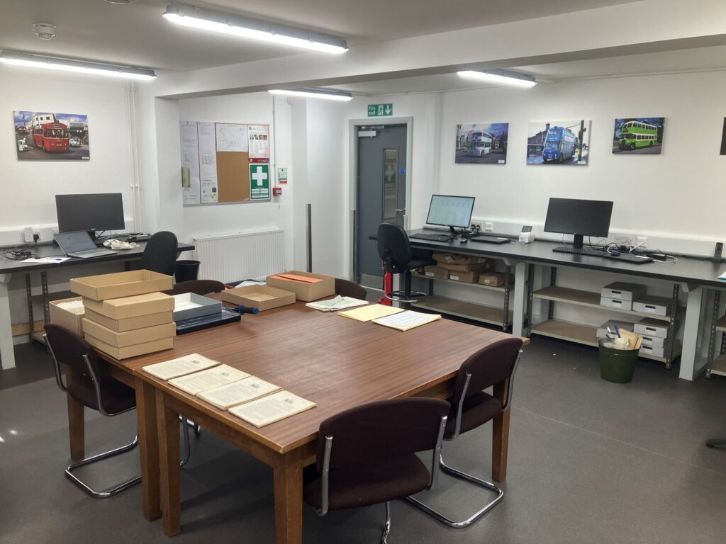 A large white room with two rectangular tables in the centre, pushed together to form one large square table. Computer desks are placed along the walls.