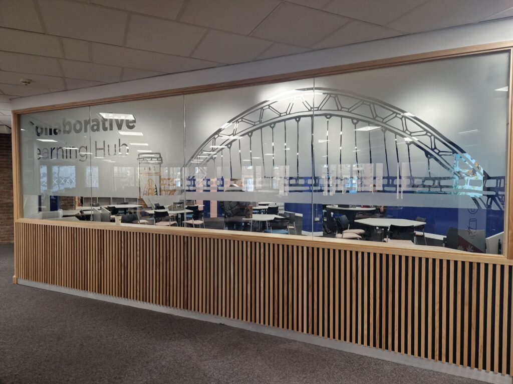 Looking through a wall containing large glass panels in the top half, into a room full of tables and chairs. Much of the glass is frosted, with a large arched bridge depicted on it, along with the words 'Collaborative Learning Hub'.