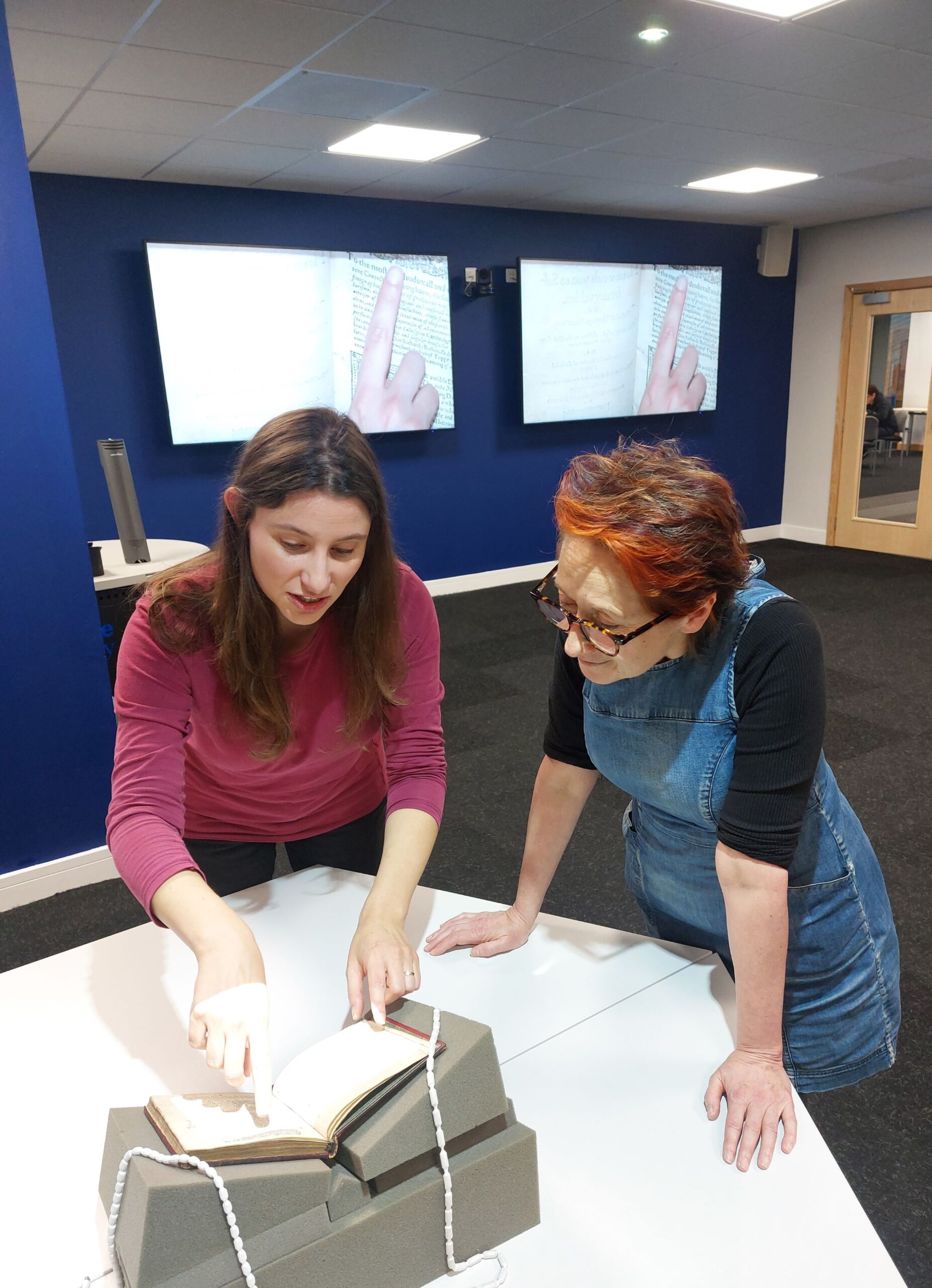 Two people stand over an open book, one of them pointing to part of a page. The part of the page that they are pointing to can be seen on two large television screens behind them.