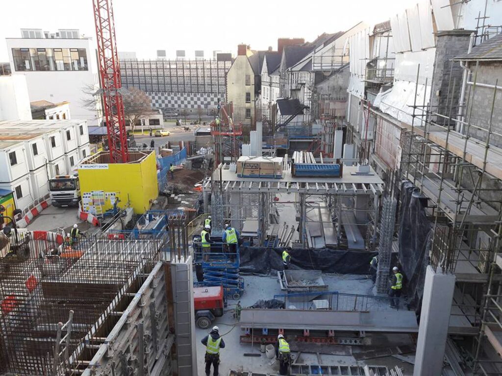 An open-air construction site. Nine construction workers are on site, at ground level.