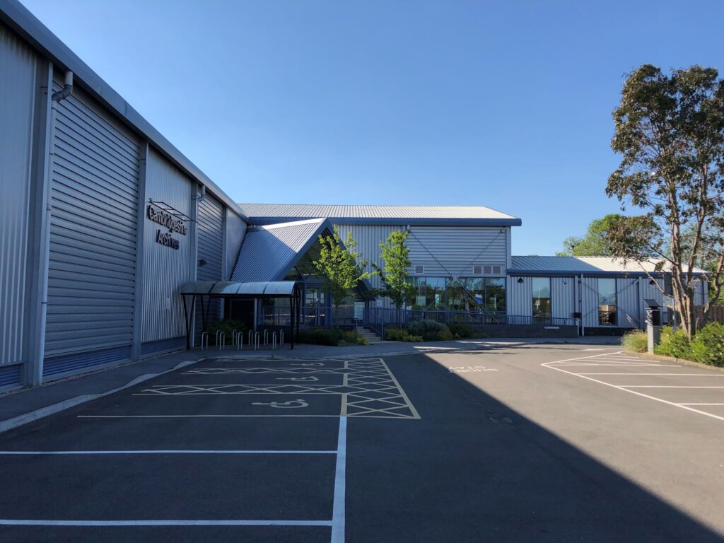 View from the car park of a large, grey, metal-clad building, which appears to be in a 'dog leg' shape. The words 'Cambridgeshire Archives' are attached to the building in large purple letters.