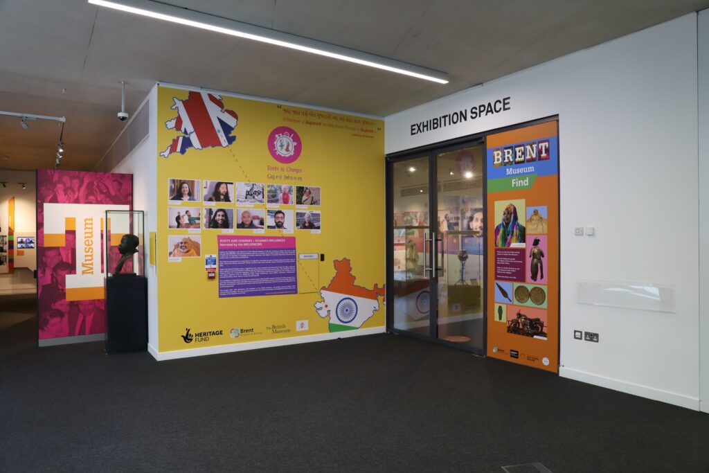 A wide open space, with wall space occupied by exhibition information, including a yellow wall with information about a project called 'Roots & Changes - Gujurati Influences'. A set of glass doors are set in one wall, with the words 'exhibition space' above in black capitals.