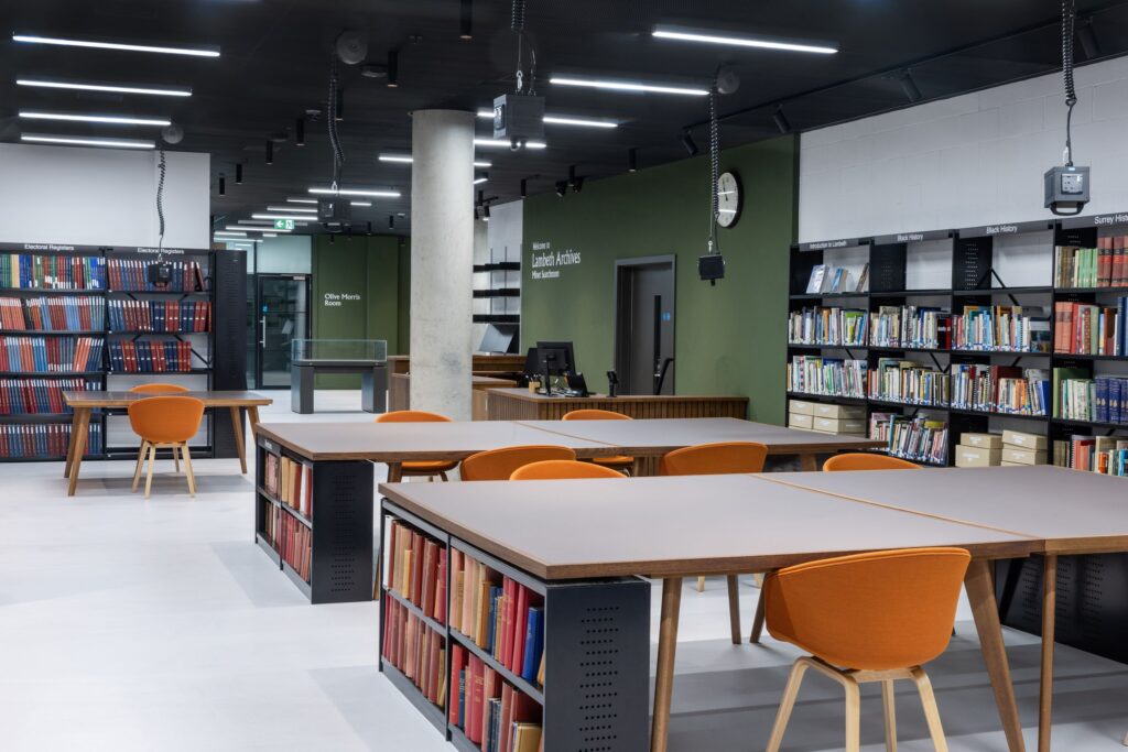 A large open space, with tall black bookshelves placed along much of the wallspace. There is a large reception desk towards the back of the space, and a few tables with orange chairs in the foreground, with seating enough for 10.