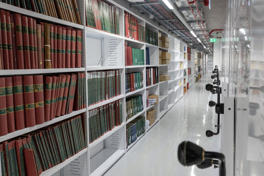 A long corridor with floor-to-ceiling shelving on the left side, full of books or cardboard archive boxes. On the other side of the corridor is a series of black three-pronged rotating handles.