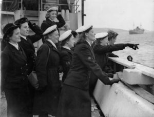 A group of women from the Women's Royal Naval Service stand on the deck of a ship looking at something on the ocean. One of the women, Jonnni Berfield, is pointing at it. 