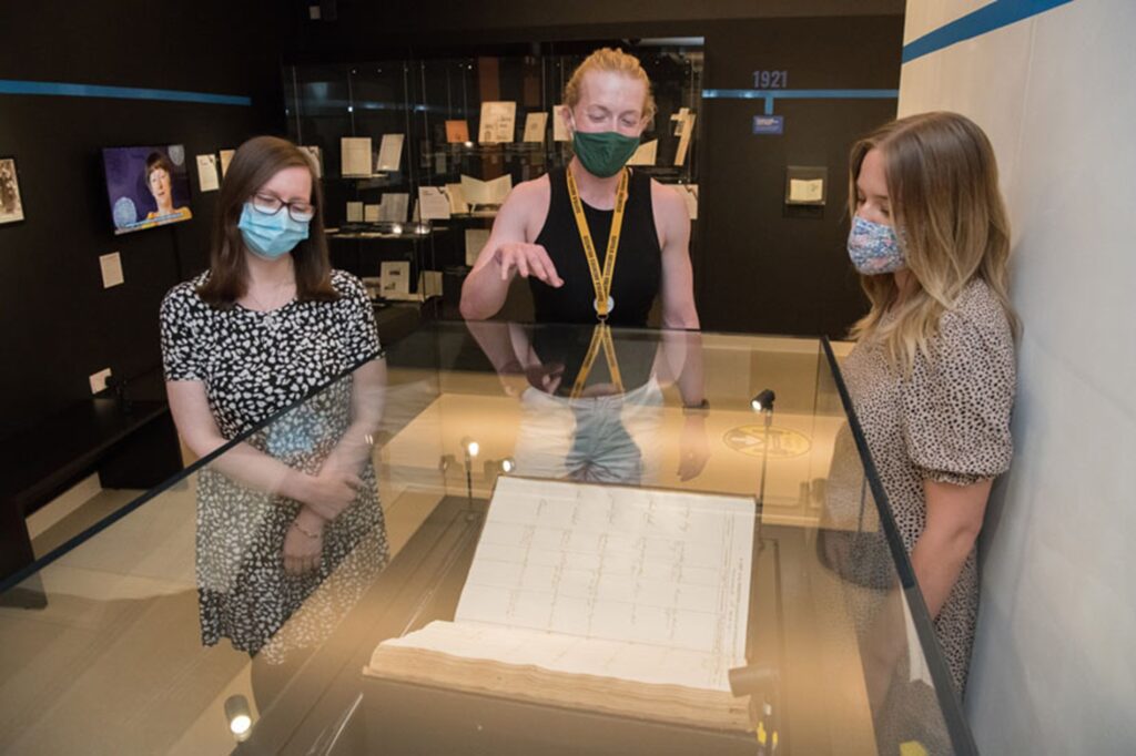 Three people stand in a room, looking at a hardback document housed in a glass display case. The document is open, and one of the people is gesturing with their right hand, as if explaining the document.