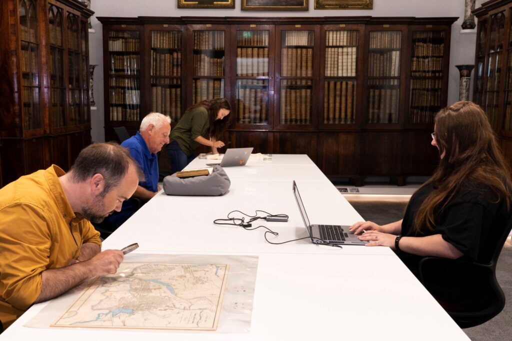 A long white table in the middle of a room, with four people around it - three are studying original documents, while the other is using a laptop. Tall antique display cabinets are around the walls of the room, filled with hardback books and documents.