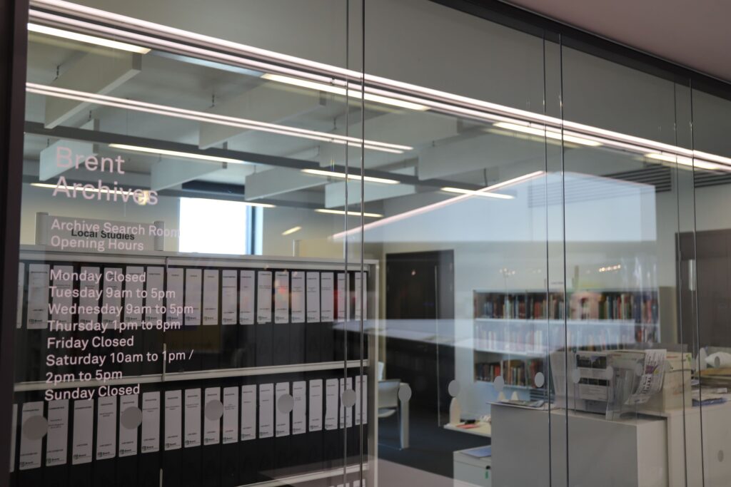 A view through glass wall dividers into a white, brightly lit room. On a far wall is a large, wide book shelf with three rows of books, while by the glass dividers there is a freestanding shelving unit containing a series of black ring binders.