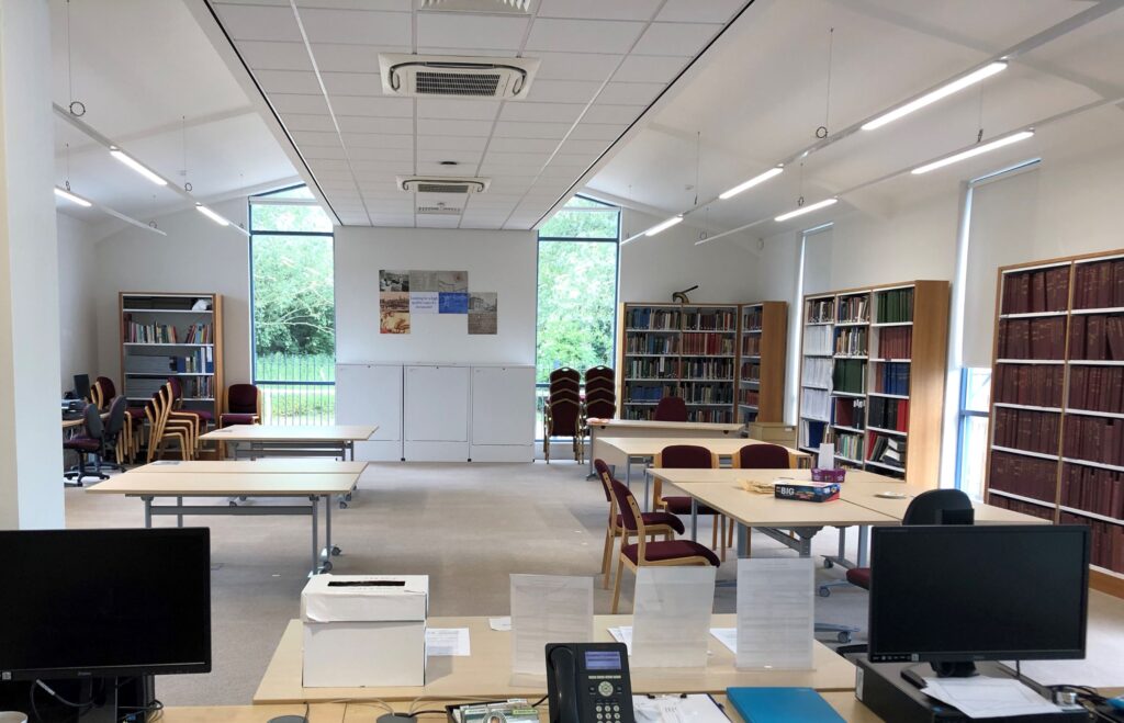 A large, open white-walled space, with tall bookshelves along the walls. Two desks with computer monitors are in the foreground; ahead of them are four large tables, one of which has chairs around it.