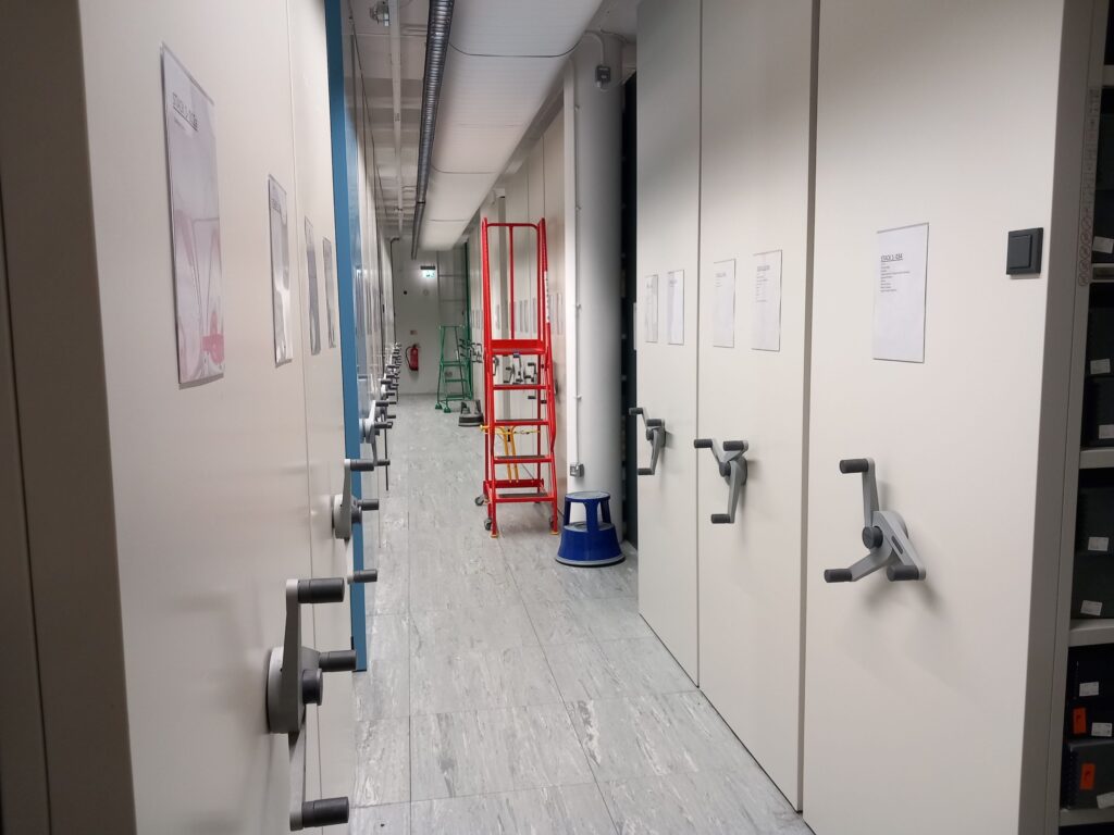 A white corridor with floor-to-ceiling racking on either side. Each row of racking has a grey three-pronged rotating handle.