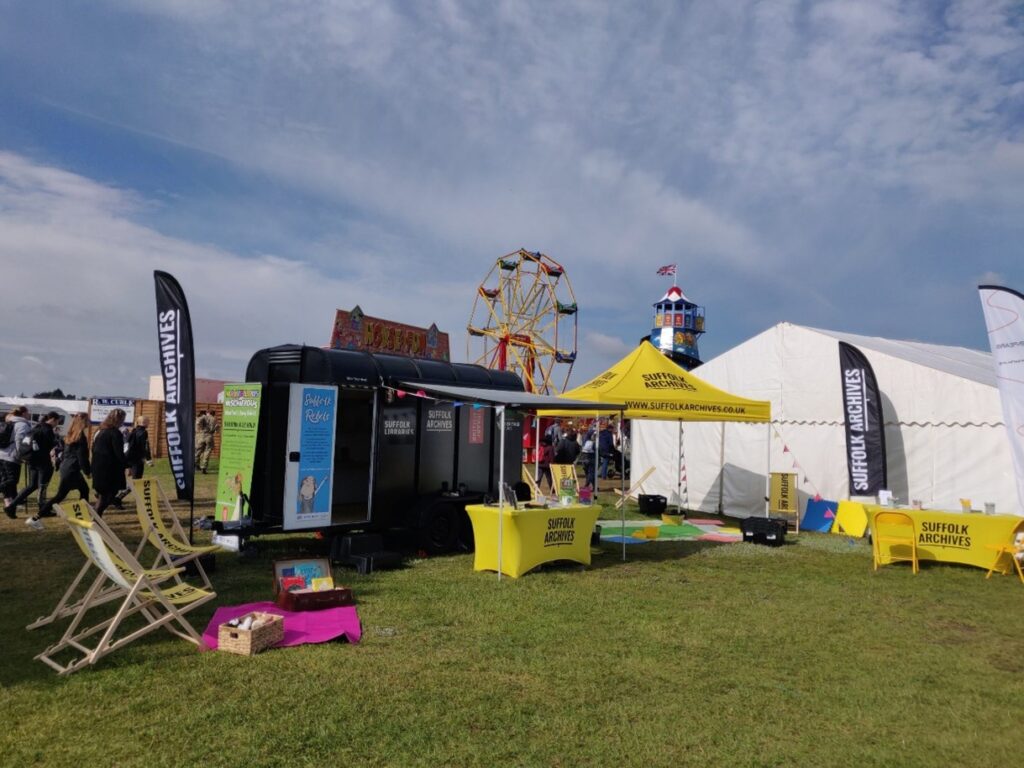 A gazebo and a tall, enclosed car trailer in a grassy field, with deck chairs and tables dotted about. A ferris wheel and helter skelter are in the background.