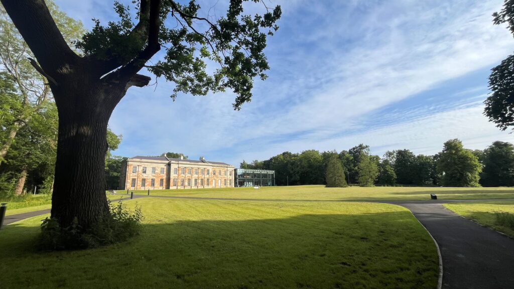 Looking past a large tree in the left foreground, across extensive lawns, on a sunny day with mainly blue skies, towards a building. The building is formed of two parts - the larger part on the left is a large orange and beige brick build, in the style of a stately home. The smaller part on the right is primarily glass, with a green frame.