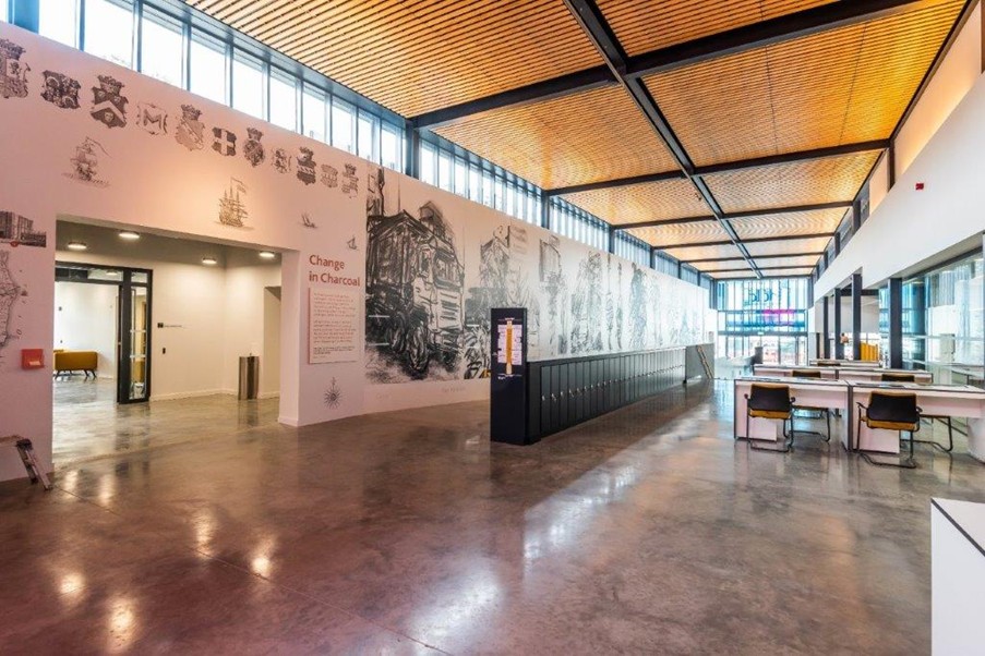 A long, open, high-ceilinged space, with bright daylight coming through high-placed windows that run along the length of the space. There is a small set of desks with chairs on the right hand side.