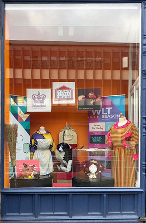 A large glass display window, housing various artefacts, including a border collie doll, two brightly coloured outfits, and a wig of red hair.