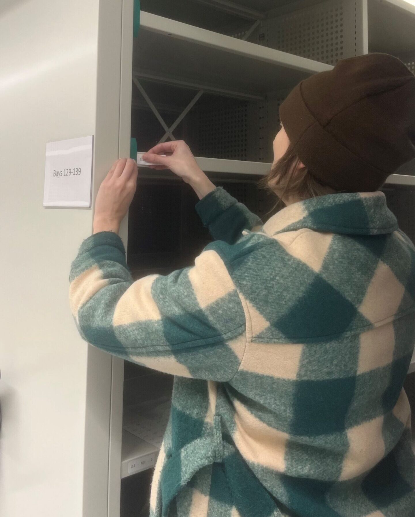 A person wearing a thick green and white checked jacket and dark brown beanie stands at some floor-to-ceiling open shelving. They are attaching a label to a shelf.