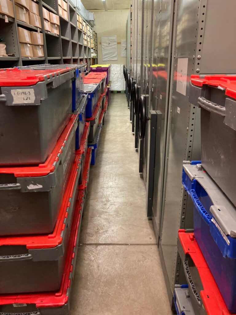 A corridor full of deep plastic boxes with folding lids, primarily grey in colour with red lids. Floor-to-ceiling open shelving is on the left, storing clusters of deep cardboard archive boxes, while the right side is taken up by floor-to-ceiling mobile racking, with black circular handles for moving racks.