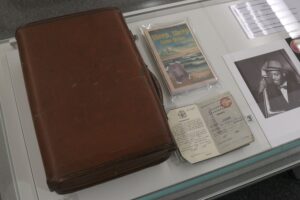 A display of several records on a white surface including a large leather bound book, a black and white picture of a man appearing to hold a double bass and an illustrated publication called 'Sheep, sheep come home'
