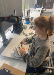 A young woman sits at a row of desks working with an open journal and a vertically mounted camera.