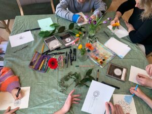 An overhead shot of table filled with flowers, leaves and pens. Several people are sat around the table creating drawings.
