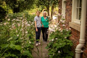 A young boy and an older woman stand by an old cobbled wall amidst dozens of flowers. They hold hands.