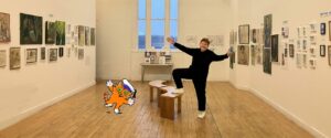 Shelia Graber stands in a large white gallery space with pictures lining the walls. Her arms are stretched out and an animated orange cat with a sailor's hat is imposed  on the image next to her pulling the same pose.