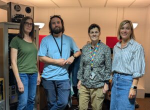The four project team members stand in a line in front of sound and video recording equipment.