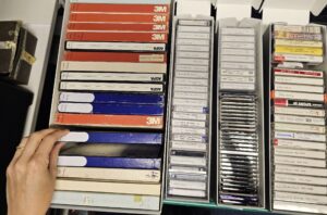 A hand flickers through boxes of labelled audio tapes and cassettes.