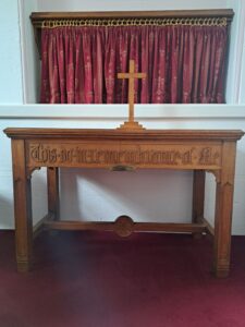 A wooden table with a wooden crucifix on it stands in front of a red and gold curtain.