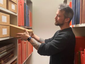 A person with arm tattoos removes a cardboard box from shelving in a repository that holds many more boxes and record books.