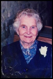 A colour image of Dorothy Fahey, taken in 2004. The photo is taken portrait style, featuring Dorothy's head and shoulders. She is smiling, wearing a royal blue shirt and navy jacket with a white rose in the lapel.