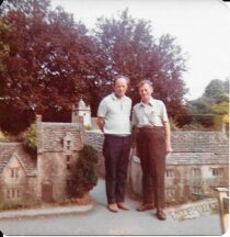 Colour photo showing Alun Mainwaring reunited with his father Daniel in 1975, 46 years after Daniel immigrated to the States. The pair stand side by side, with a model village in the background.