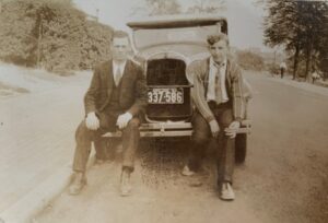 Sepia photo of Daniel and his friend, Evans 'Jack' Griffiths, in America, 1931. They are both sitting on the bumper of a car that is parked by the side of the road.
