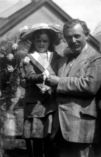 A black and white photo of David Lloyd George with his daughter, Megan, in 1911. The pair are standing shoulder to shoulder, in contemporary suits, in a garden. Megan is wearing a hat and holding a trowel.