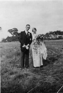 Black and white photo of Susan May and Reginald on their wedding day. The pair are standing in a grassy field. Susan May has one hand through Reginald's arm, while her other holds a large bouquet.