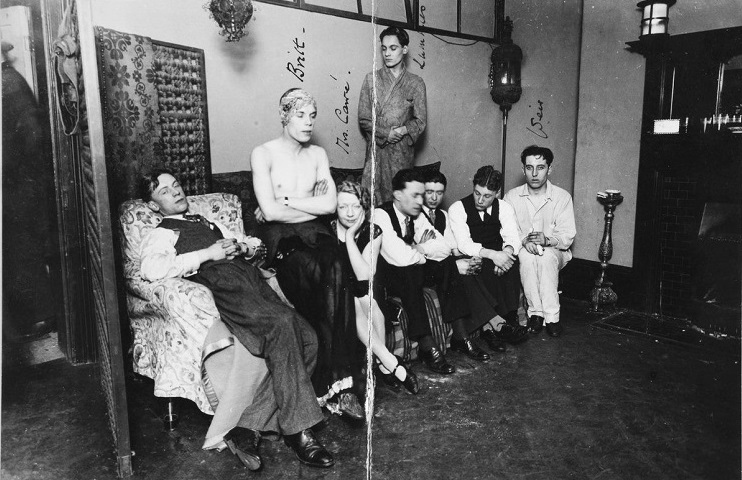 Robert Britt (Bobby, second left) with friends. Bobby is seated on the arm of a sofa, wearing a headdress and a dress, which has been removed from his chest, leaving his chest exposed. A friend sits in a chair next to him, and five other friends on the sofa. Constance Carre is on his right, her head supported by her hand. The photo is taken from the police raid on Fitzroy Square, used as evidence. (catalogue ref: CRIM 1/387)